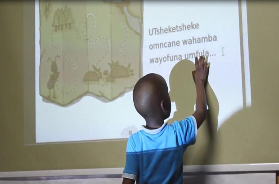 https://youtu.be/8fccqag954i: A child in Emily Mbhele s class reading a storybook page projected on to the classroom wall.